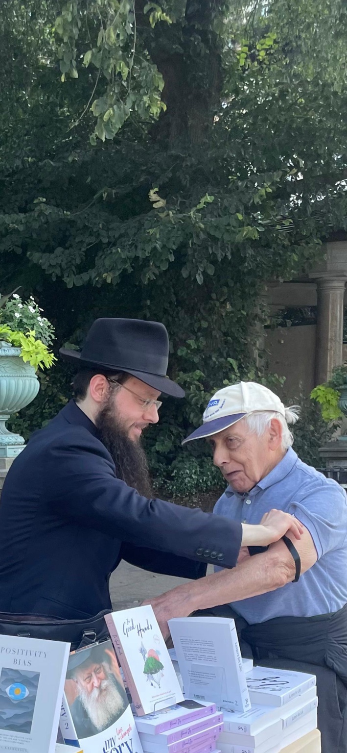 Rabbi Yisroel Tewel helping a man put on tefillin beside a table of Jewish books.